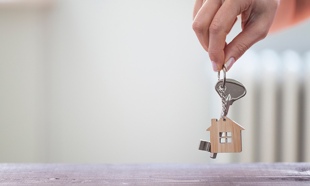 Woman hand holding key with house shaped keychain. Mortgage concept. Natural light room interior. Real estate, moving home or renting property.