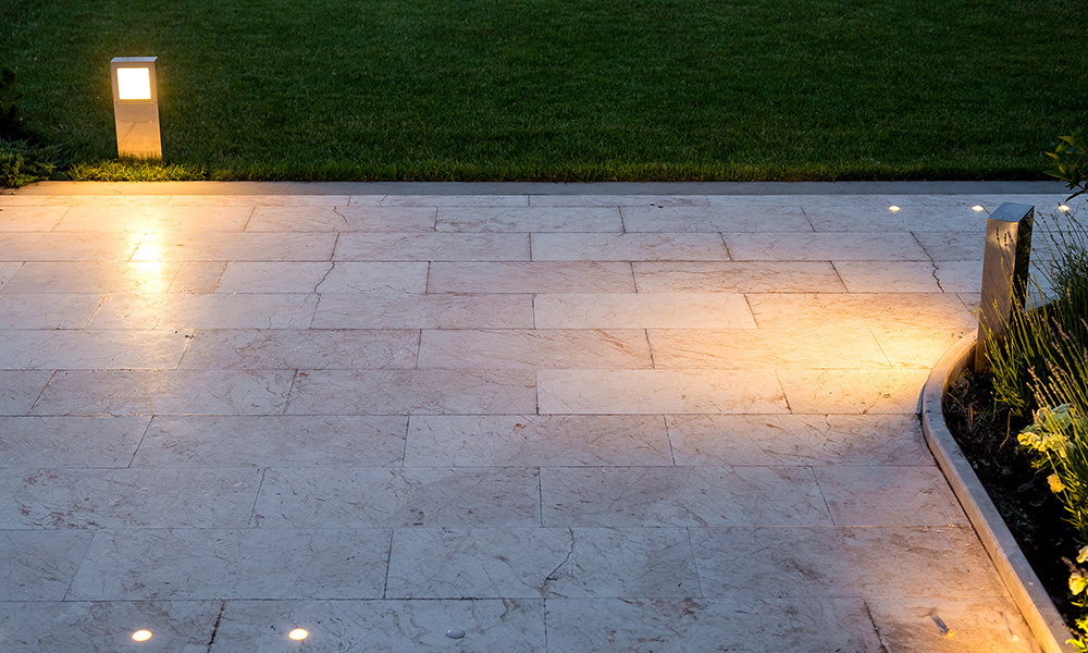 The pavement of marble tiles and a stone border area in the evening illuminated by ground lanterns in a metal housing shining a warm light around the lawn.